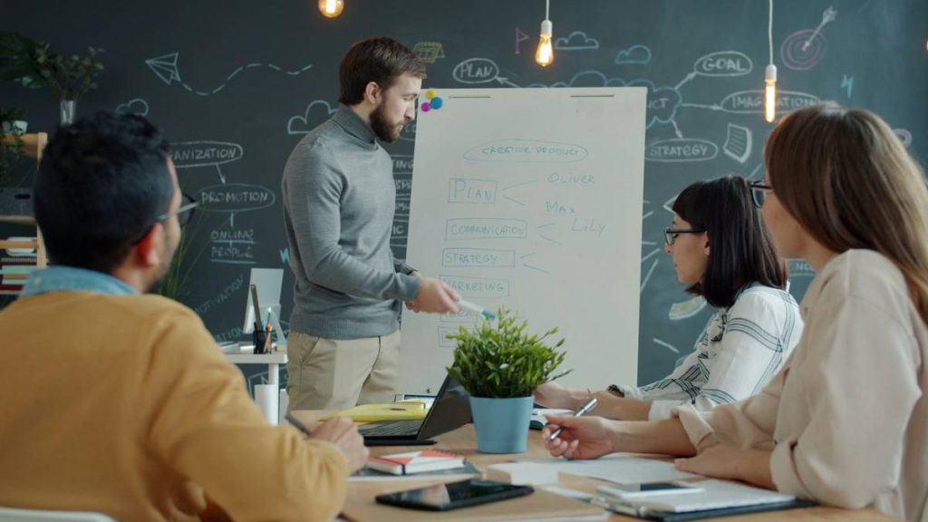 Office worker is making presentation to colleagues talking writing on whiteboard discussing marketing strategy. People and business discussions concept.